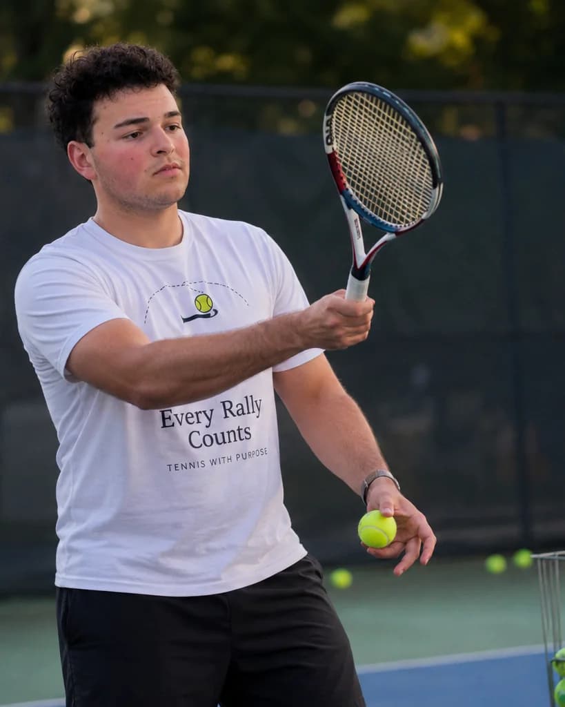 Jonah making contact on a forehand, ball in hand, on a public tennis court.